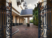 Ornate wrought iron gates open to a brick driveway leading to a covered entrance of a beige building with large pillars and lush greenery on the right.