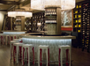 Modern bar area with white stools, illuminated counters, wine bottles on shelves, and large crystal chandeliers overhead.