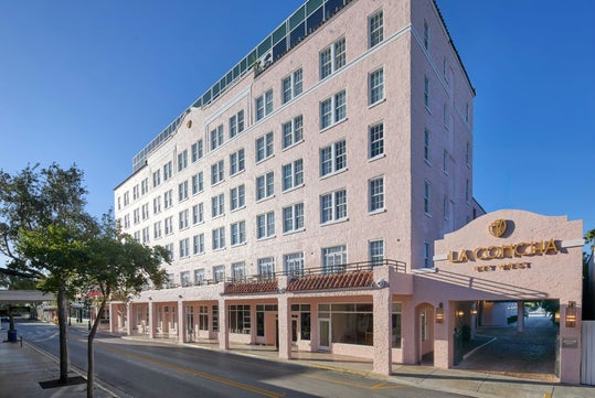 A pink, six-story hotel building labeled "La Concha Key West" stands on a quiet street under a clear blue sky.