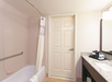 Bathroom with a shower-tub combo, curtain, beige door, wall-mounted grab bar, granite countertop with sink, mirror, and black coffee maker on the right.