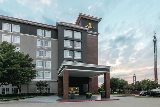 Exterior view of a multi-story hotel with a sign reading "La Quinta." The building features large windows, a covered entrance, and is surrounded by trees and a parking area.