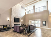 Modern lobby with high ceilings, large windows, a stone fireplace, a wall-mounted TV, seating area with purple chairs and dark sofas, and a patterned rug on tiled floor.