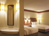 Hotel room with two double beds, a nightstand, and wall-mounted lamps. A small mirrored area with a countertop is visible on the left. Neutral color scheme with beige walls and bedding.