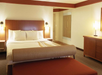 A hotel room with a large bed, two bedside lamps, a bench at the foot of the bed, and a dresser. Walls are light-colored, and the carpet is brown.