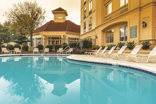 Outdoor swimming pool surrounded by white loungers and greenery, adjacent to a beige building with large windows and a red-roofed structure.