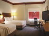Hotel room with two double beds, a TV on a wooden dresser, an office chair at a desk, and a window with a red curtain featuring a leaf design.