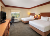 Hotel room with two double beds, a desk with a chair, a flat-screen TV, and a window with a view of trees. The room has beige walls and carpeted floor.