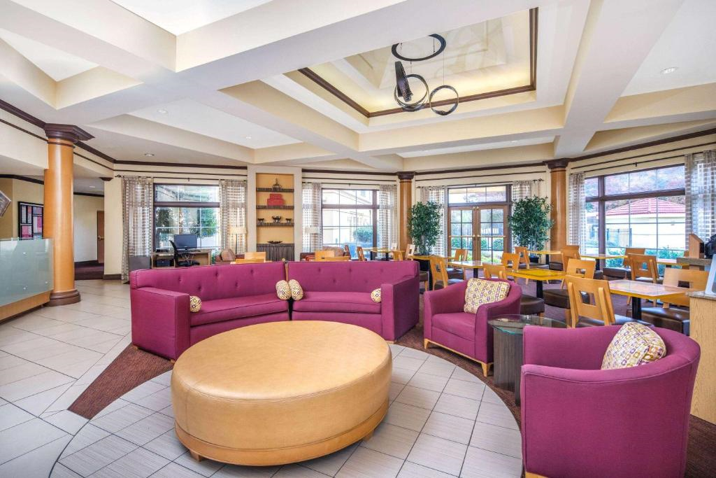 Spacious hotel lobby with purple sofas and a large round ottoman, surrounded by tables and chairs, featuring modern ceiling lights and large windows.