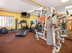Small gym with exercise equipment including a treadmill, elliptical, stationary bikes, and a weight machine. Carpeted floor, yellow and orange walls, wall-mounted TV.