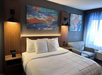 A hotel room with a neatly made bed, abstract artwork above the headboard, two wall sconces, a small couch, and a window with curtains.