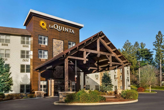 Front view of a La Quinta hotel with a wooden canopy entrance, surrounded by trees under a clear blue sky.
