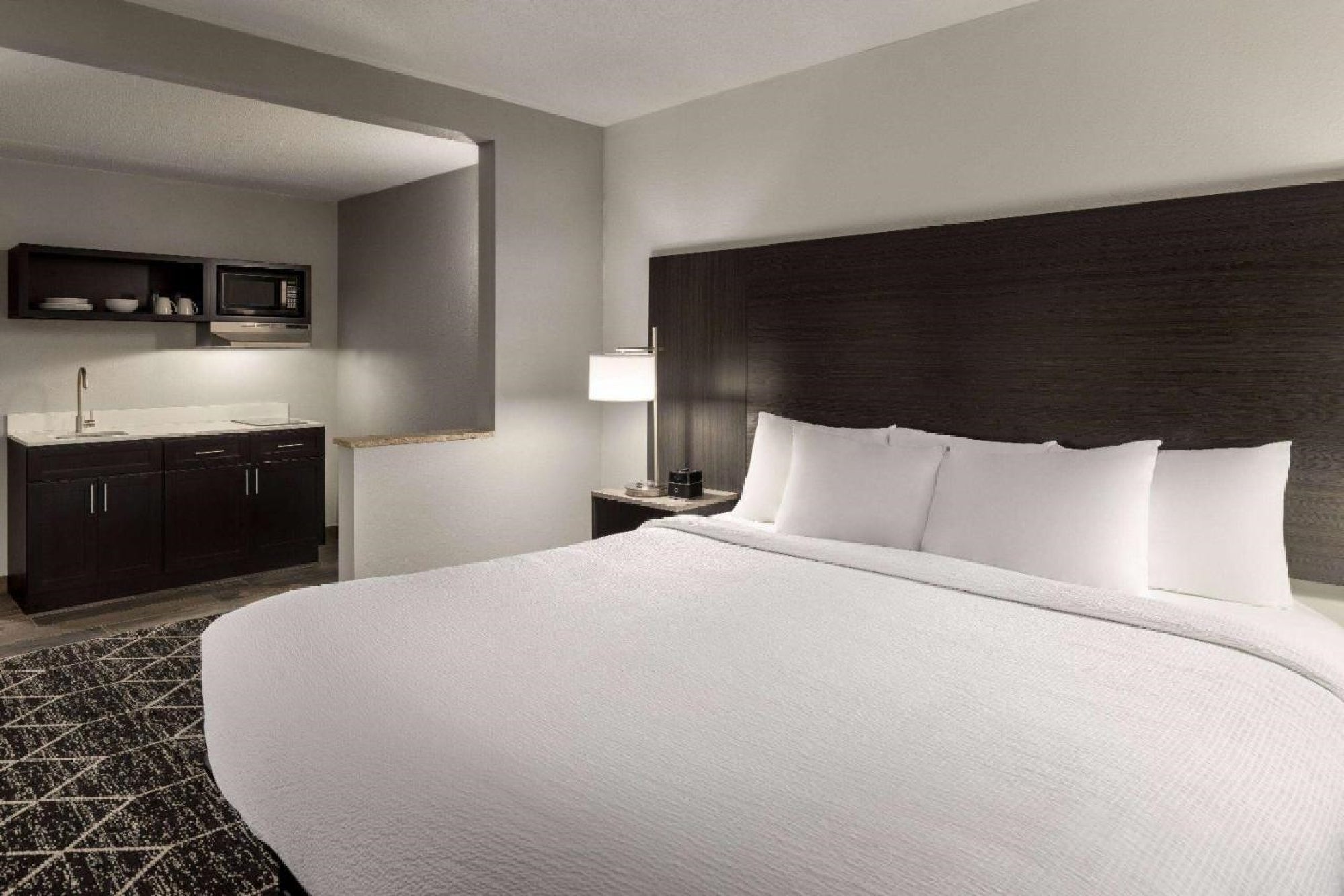 Hotel room with a king-sized bed, dark wooden headboard, and side lamp. A kitchenette with a sink, microwave, and cabinets is visible in the background.