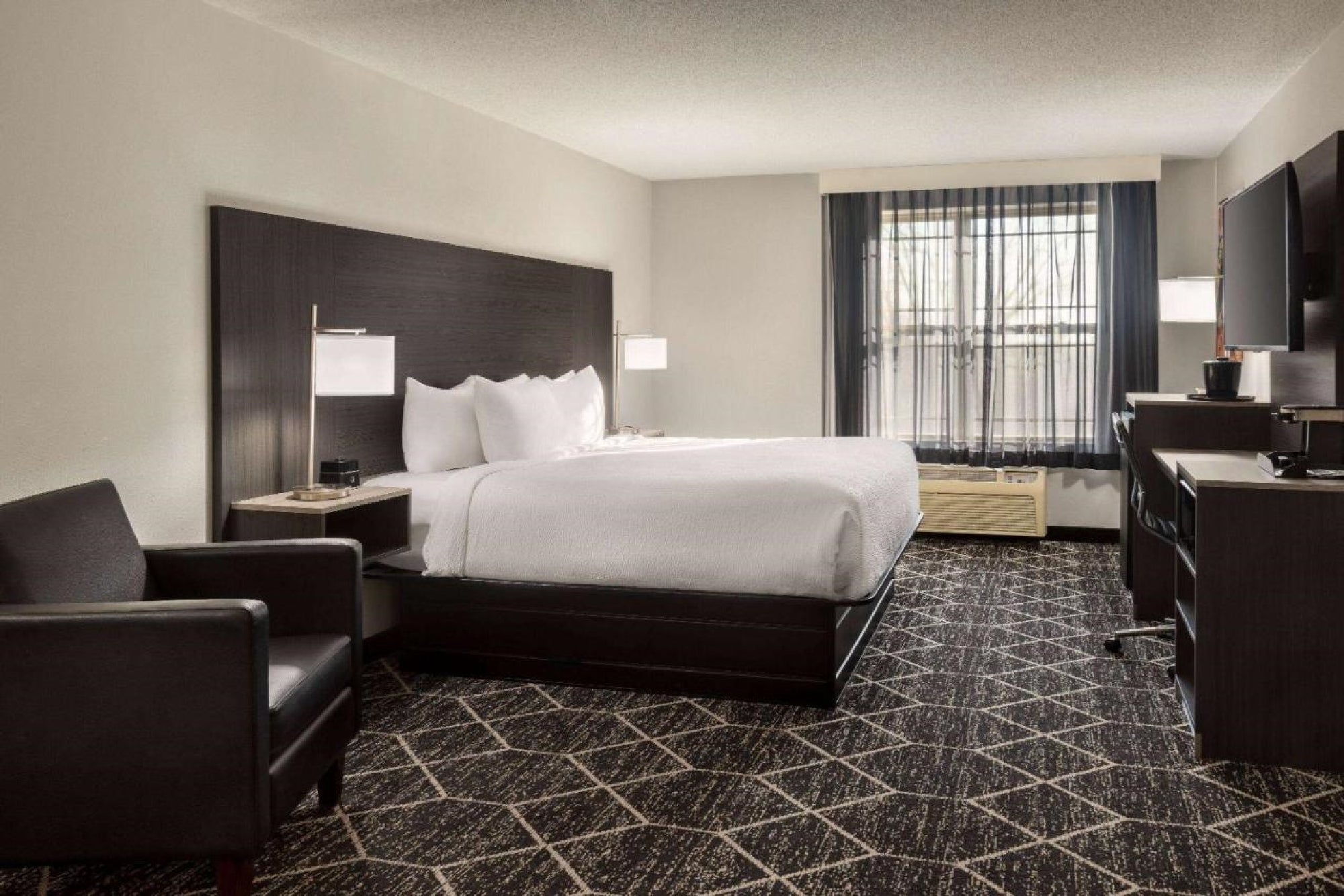 Hotel room with a king-sized bed, two bedside lamps, an armchair, a desk with a TV, and a window with sheer curtains. The carpet has a geometric pattern.
