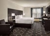 Hotel room with a king-sized bed, two bedside lamps, an armchair, a desk with a TV, and a window with sheer curtains. The carpet has a geometric pattern.