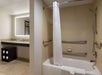 Hotel bathroom with a bathtub, shower curtain, and towel. Sink and mirror are visible to the left. Grab bars are installed around the tub.