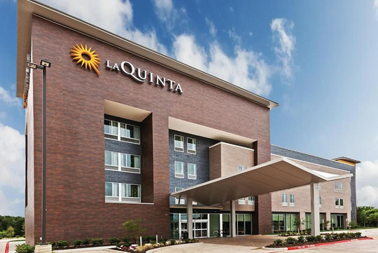 Exterior view of a La Quinta hotel with a large sign, multiple windows, and a covered driveway at the entrance under a partly cloudy sky.