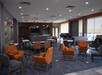 Modern lounge area with blue carpet, orange and gray chairs, small tables, and large windows letting in natural light. The space has a contemporary design and ceiling lighting.