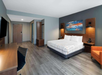 Modern hotel room with a king-size bed, desk, TV, orange chair, wood floors, and wall art above the bed.