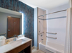 Accessible hotel bathroom with bathtub, grab bars, shower curtain, large mirror, sink counter, towels, and toiletries. Door is closed; lighting is bright and even.