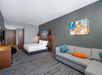 Modern hotel room with a bed, desk, wall-mounted TV, gray sofa with colorful cushions, and abstract artwork on the walls.
