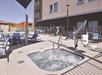 Outdoor hotel area with a small jacuzzi in the foreground and a larger pool in the background. Several lounge chairs and a table with chairs are arranged around the area.