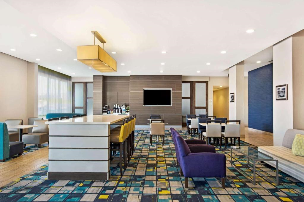 Modern hotel lobby with a bar counter, seating area with purple and gray chairs, patterned carpet, and a wall-mounted TV.