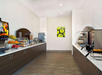 A clean, modern buffet area with fruit, bread, pastries, beverages, and a waffle maker. White walls, wooden cabinets, and a framed picture of green apples decorate the space.