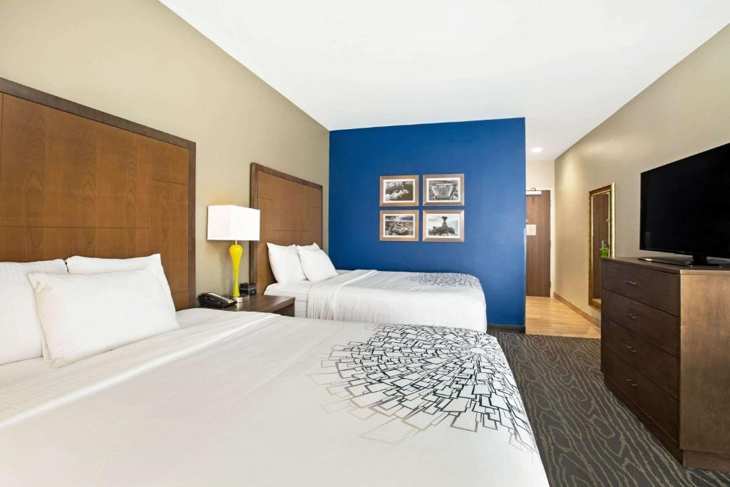 Hotel room with two beds, a nightstand with a lamp, a blue accent wall with framed pictures, a dresser with a TV, and a hallway leading to the entrance.