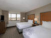 Hotel room with two double beds, a window view, TV on a dresser, lamps, and neutral decor.