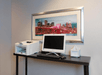 A computer and printer sit on a dark table beneath a framed cityscape photo.