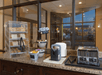 Hotel breakfast setup with bread, bagels in a plastic case, muffins on tiers, a toaster, and condiments on a counter. Multiple windows in the background.