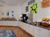 Hotel breakfast area with buffet stations, coffee and juice dispensers, a fruit basket, and wall art.