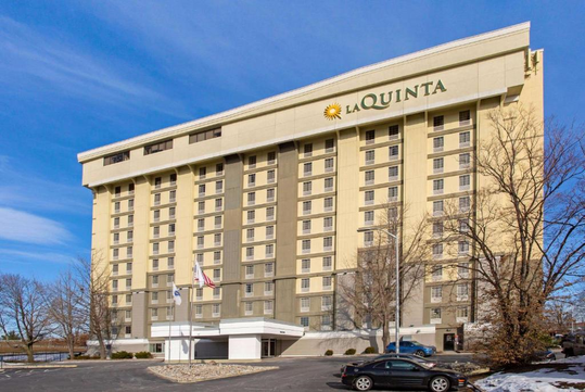 Large hotel with "La Quinta" signage, featuring multiple floors, beige and gray facade, surrounded by trees and a clear blue sky.