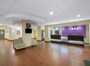 Hotel lobby with wood flooring, two armchairs, a wall-mounted TV, and a reception desk with purple accent wall. Ceiling lights and decorative panel above the TV.
