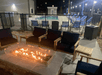 An outdoor fire pit in the middle of the several wooden chairs with black cushions and an outdoor swimming pool in the background.