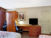 Hotel room with a desk, office chair, and TV. A wardrobe and framed picture are on the left, and two bed edges are visible. A door is on the right.