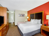 Hotel room with a large bed, wooden headboard, and red accent wall. There's a nightstand with a lamp, a dresser with a TV, and framed art on the wall. Hallway and entrance are visible.