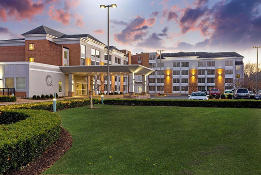Hotel building with a modern design, well-lit entrance, and surrounding greenery; several cars are parked in front. The sky is colorful during sunset.