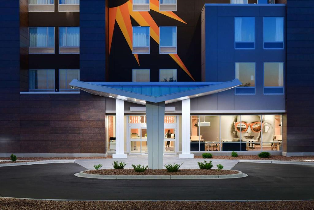 Modern hotel entrance with a large canopy, surrounded by glass windows. The facade features a geometric sun design and an image of a face with sunglasses. Small landscaped area in front.
