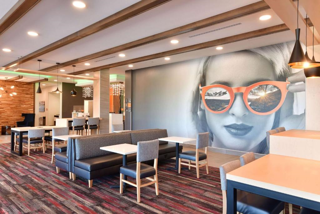 Modern cafe interior with a large wall mural of a woman wearing orange sunglasses. Tables and chairs are arranged neatly, and pendant lights hang from the ceiling.