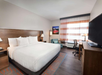 Hotel room with a large bed, armchair, and desk. A window with striped curtains provides natural light. There’s a wall-mounted TV opposite the bed and a piece of artwork on the wall.