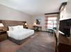 Hotel room with a king-sized bed, desk and chair, armchair, large TV, and window with curtains. Neutral tones and modern decor.