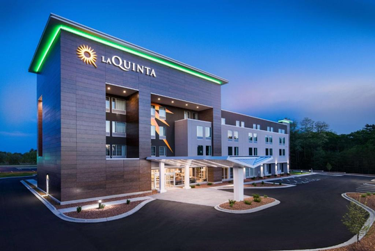 A modern La Quinta hotel building with illuminated signage, surrounded by a parking area, against a dusk sky.
