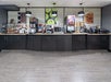 Hotel breakfast buffet area with a counter displaying a microwave, beverage dispensers, baskets of bagels, cereal, and baked goods, with food posters on the wall behind.