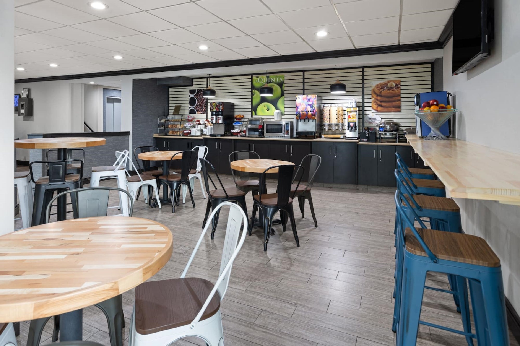 Modern self-service breakfast area with wooden tables, metal chairs, bar seating, and a counter offering food, drinks, and fruit under bright ceiling lights.