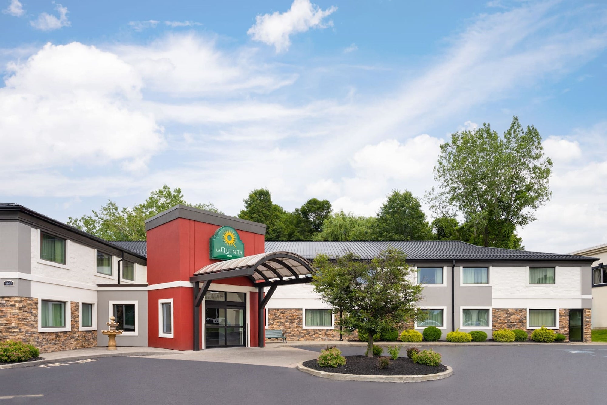 Exterior view of a two-story La Quinta Inn & Suites hotel with a red entrance canopy, surrounded by greenery and a paved driveway under a partly cloudy sky.