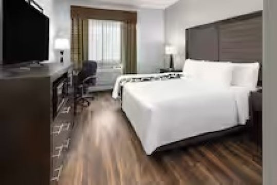 A hotel room with a queen bed, white bedding, wooden floor, desk with chair, dresser, TV, and window with curtains.