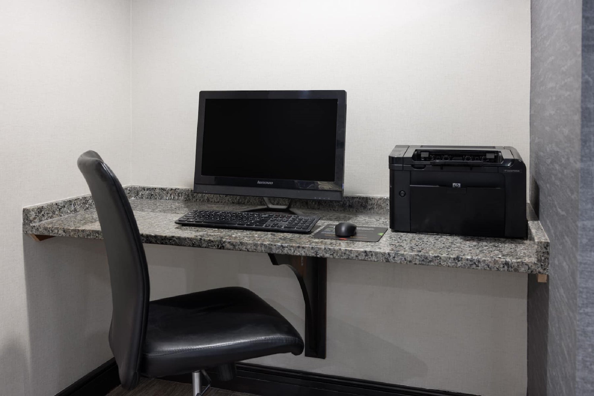 A small office corner with a desktop computer, keyboard, mouse, and a printer on a granite countertop, next to a black office chair.