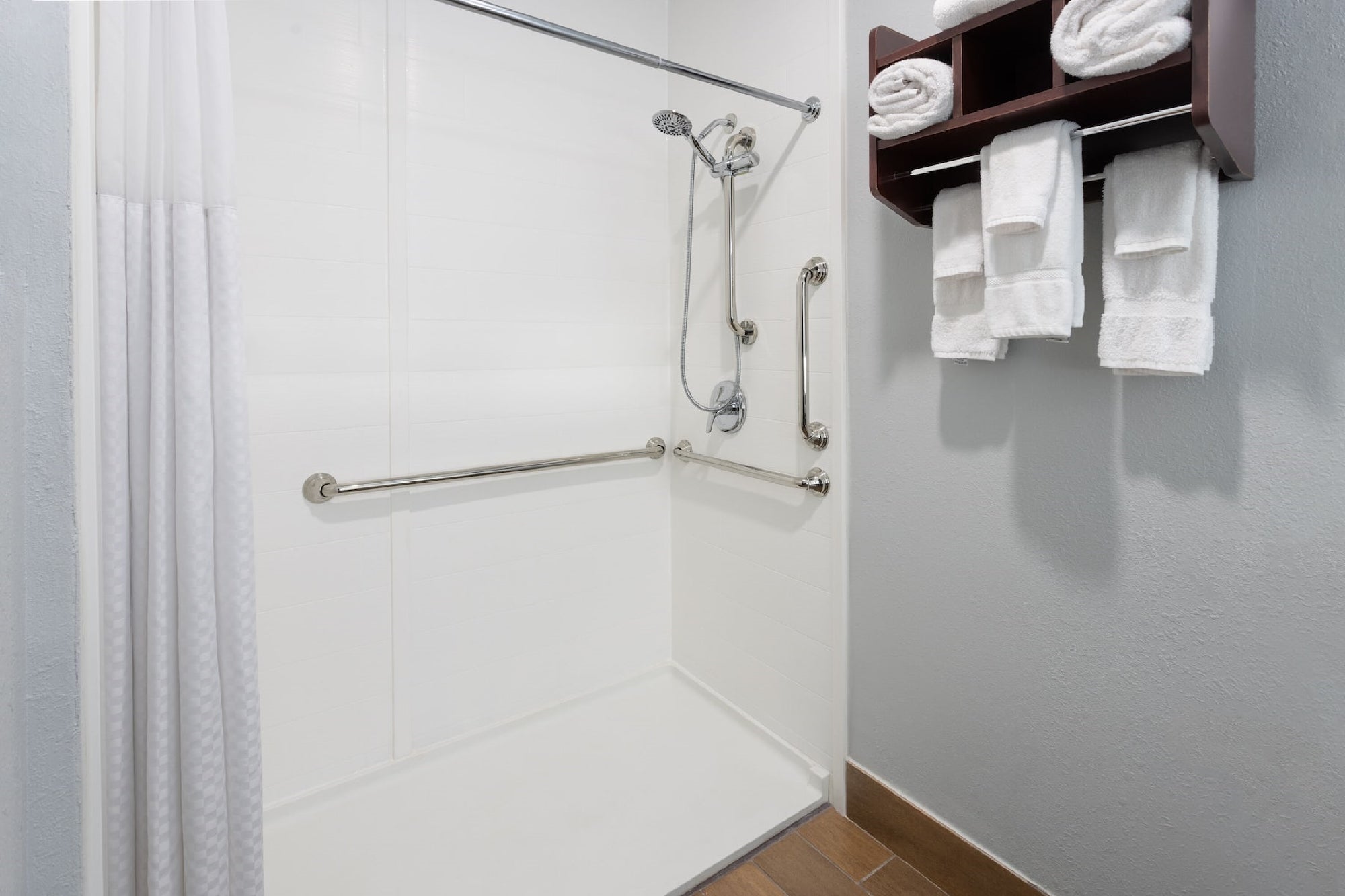 Accessible walk-in shower with grab bars, handheld showerhead, and a curtain. Wall-mounted shelves hold rolled towels and folded washcloths nearby.