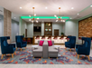 Modern lobby with blue and gray seating, colorful rug, a bar with backlit shelves, and decorative lighting fixtures.